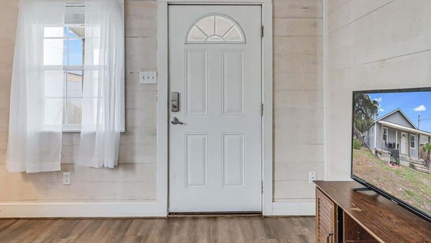 A room with a white door, a window with sheer curtains, a wooden floor, and a television displaying a house.