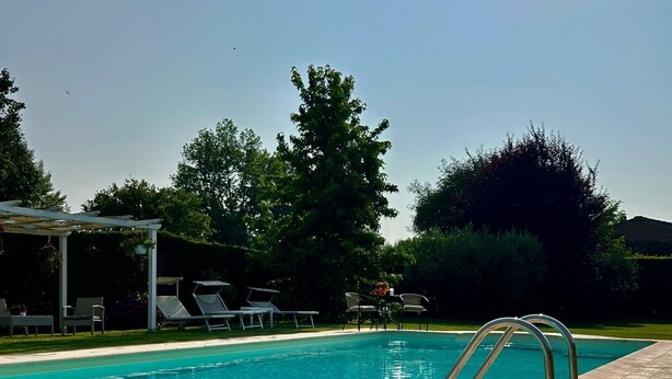 En swimmingpool med kristallklart blått vatten, omgiven av en kakelbeklädd terrass, och en pergola med stolar.