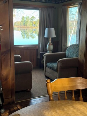A cozy room with a wooden table, two armchairs, a lamp, and a view of a lake through a window. - Beavers Resort - Cabin 4 (Mercer)