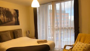 A hotel room with a large bed, a chair with a yellow blanket, a painting on the wall, and a view through sheer curtains. - 1 bed & 1 bath apartment large balcony - 10 min walk from new street station (Birmingham)