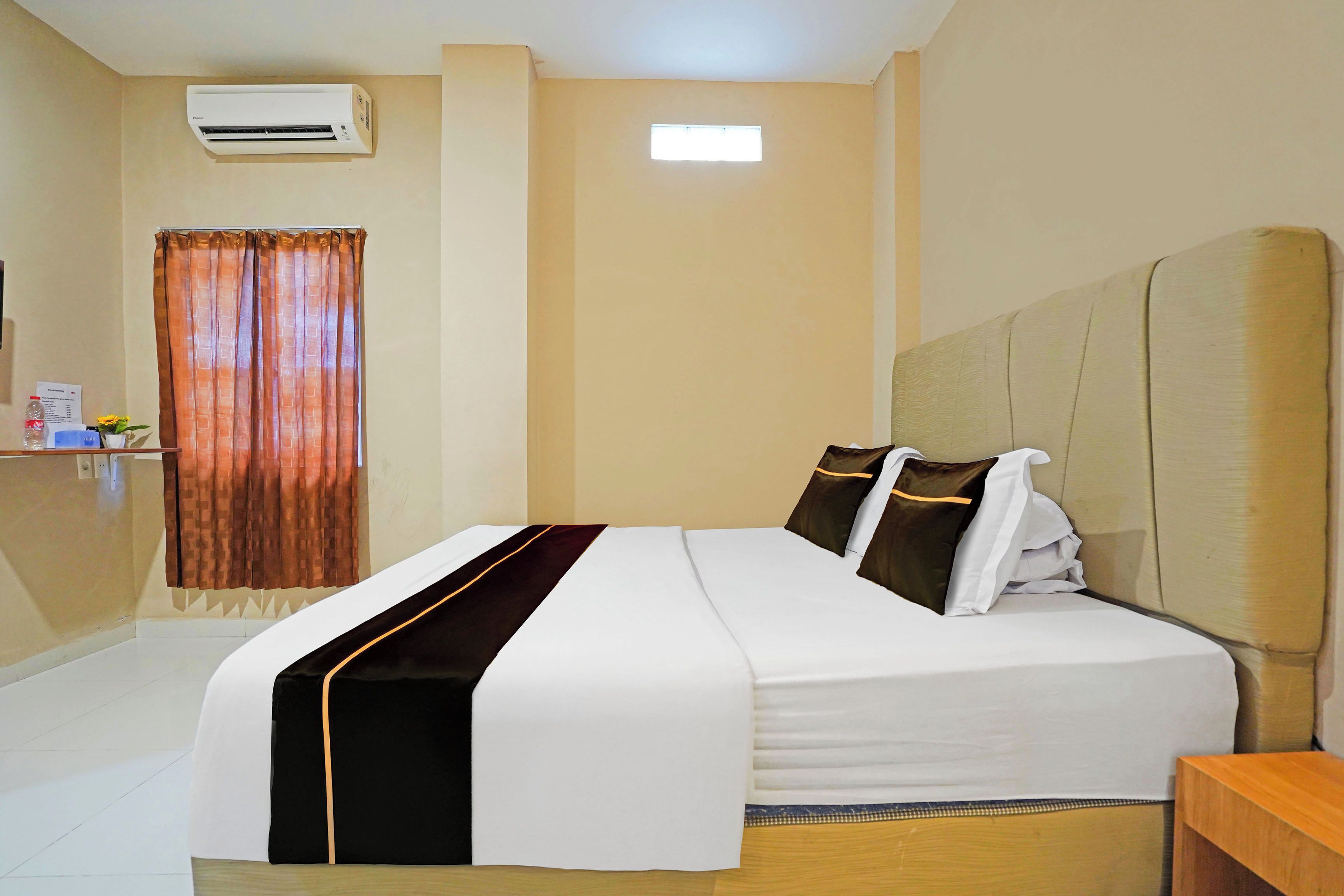 Quarto de hotel com cama, mesa de cabeceira, janela com cortinas, ar condicionado e uma pequena mesa com uma garrafa e um vaso.