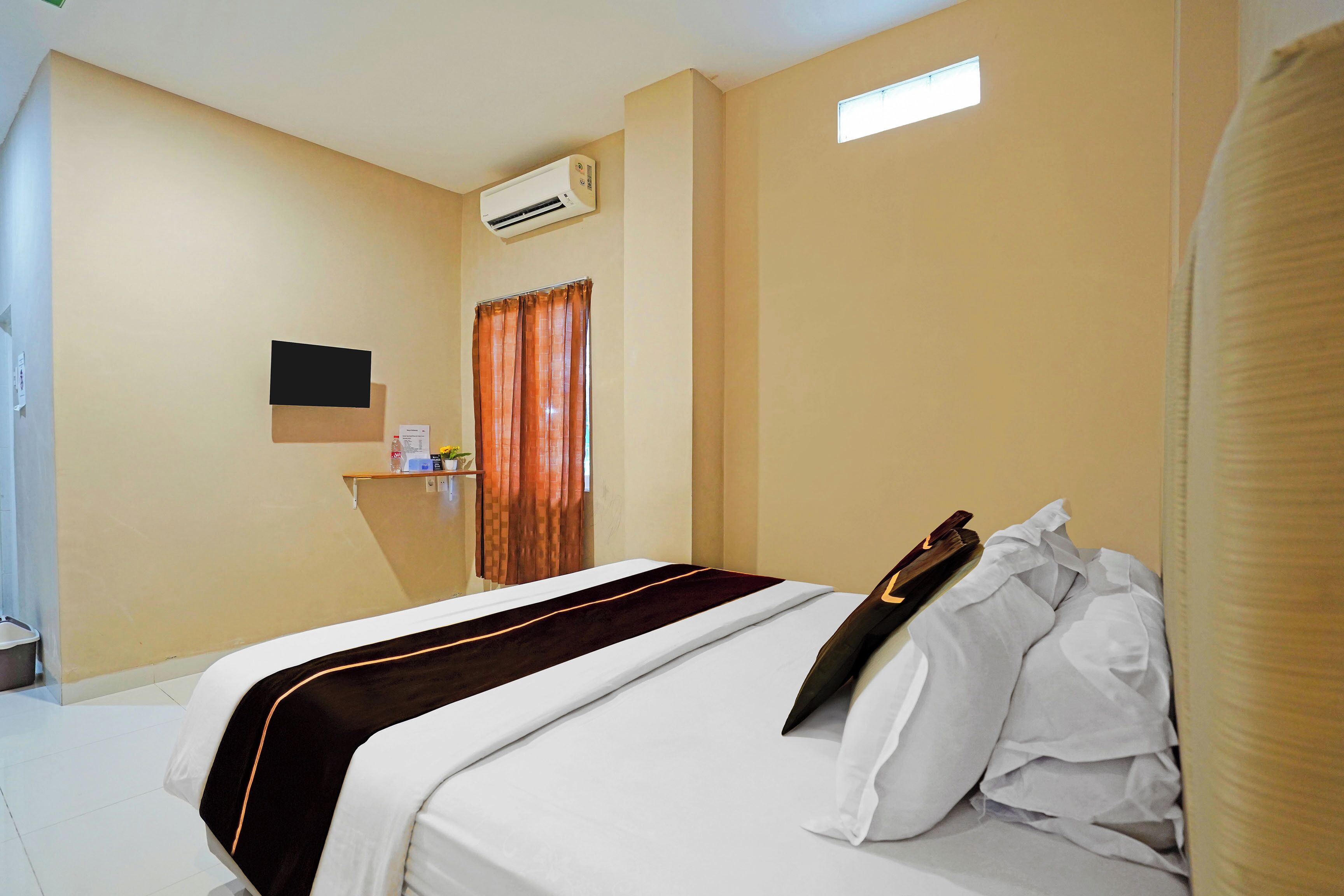 A hotel room with a bed, a flat-screen TV mounted on the wall, an air conditioning unit, and a window with curtains.