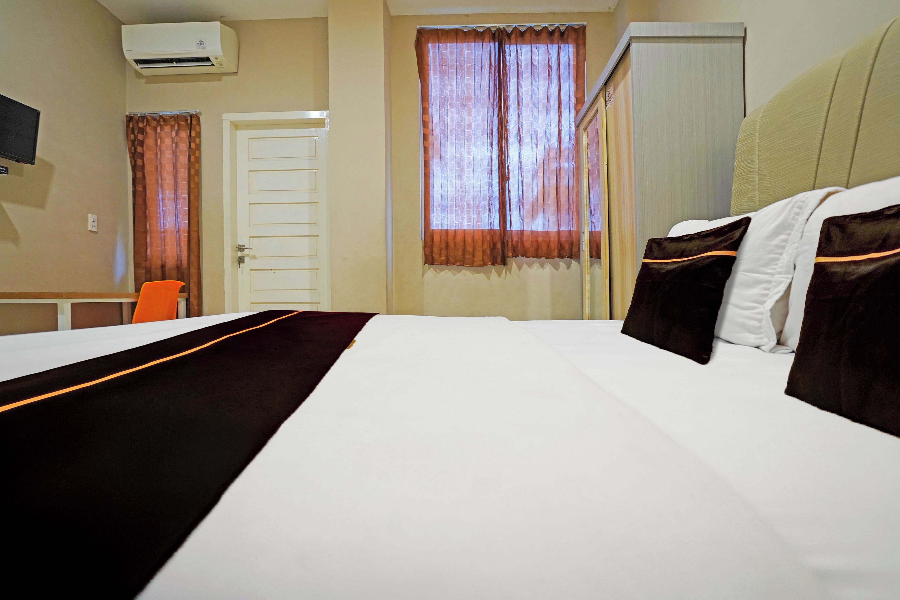 A hotel room with a bed, a desk, an orange chair, a window with curtains, and an air conditioning unit.