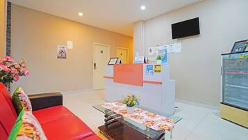 A reception area with a red couch, a glass-top table with a floral tablecloth, a reception desk, a wall-mounted TV, and a refrigerator.