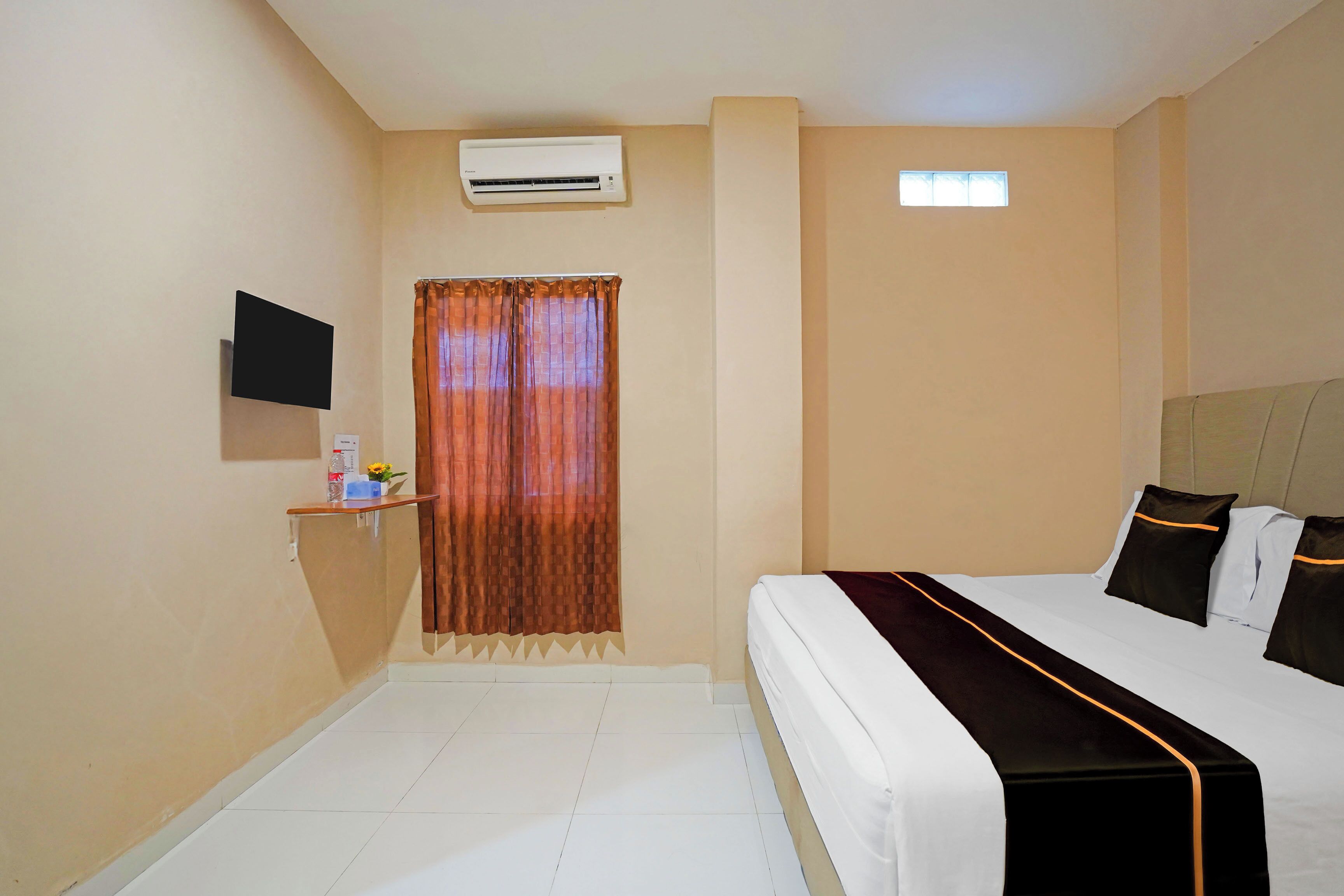 A hotel room with a bed, a TV, an air conditioner, a window with curtains, and a small shelf with items.