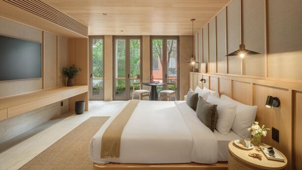 A modern bedroom with a large bed, a TV, a dining area with chairs, and a view through large windows.