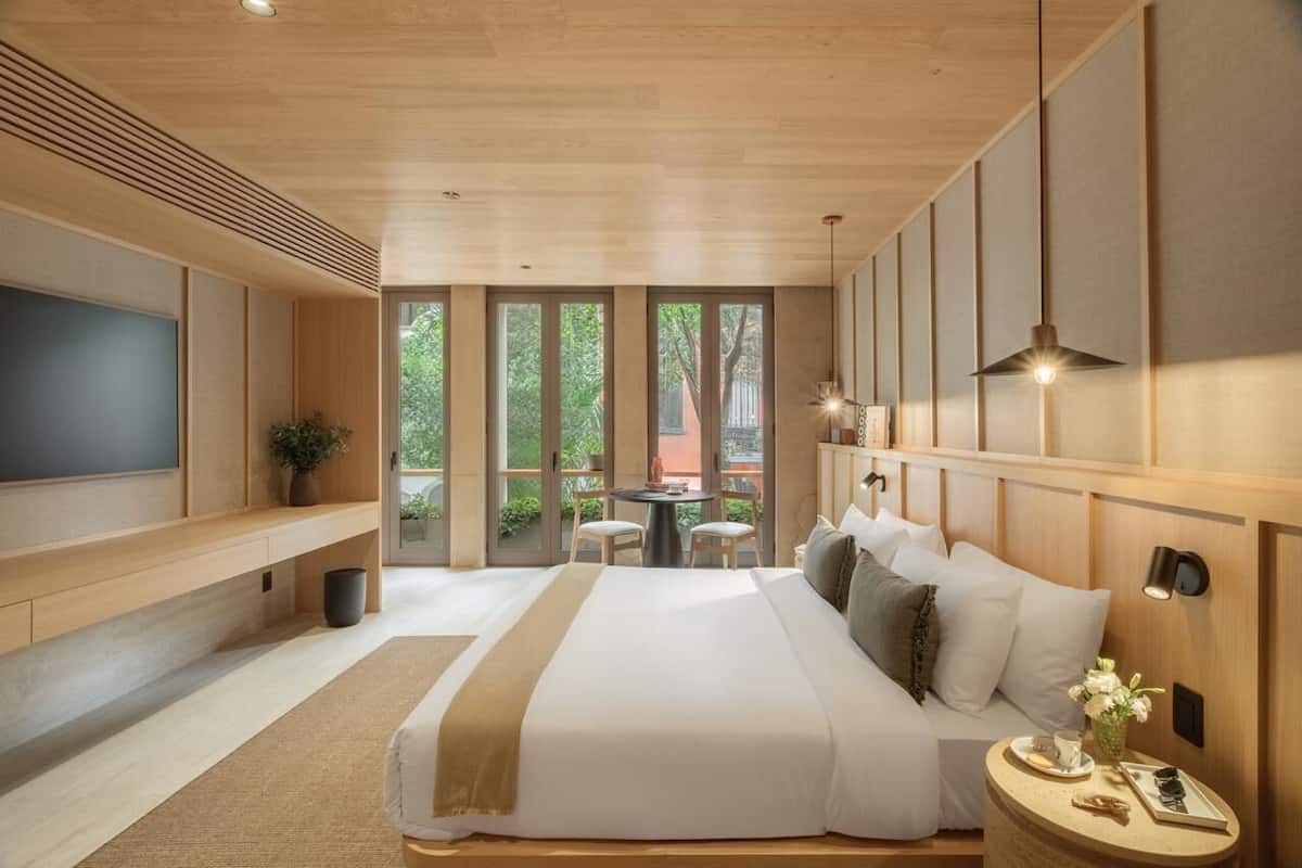 A modern bedroom with a large bed, a TV, a dining area with chairs, and a view through large windows.