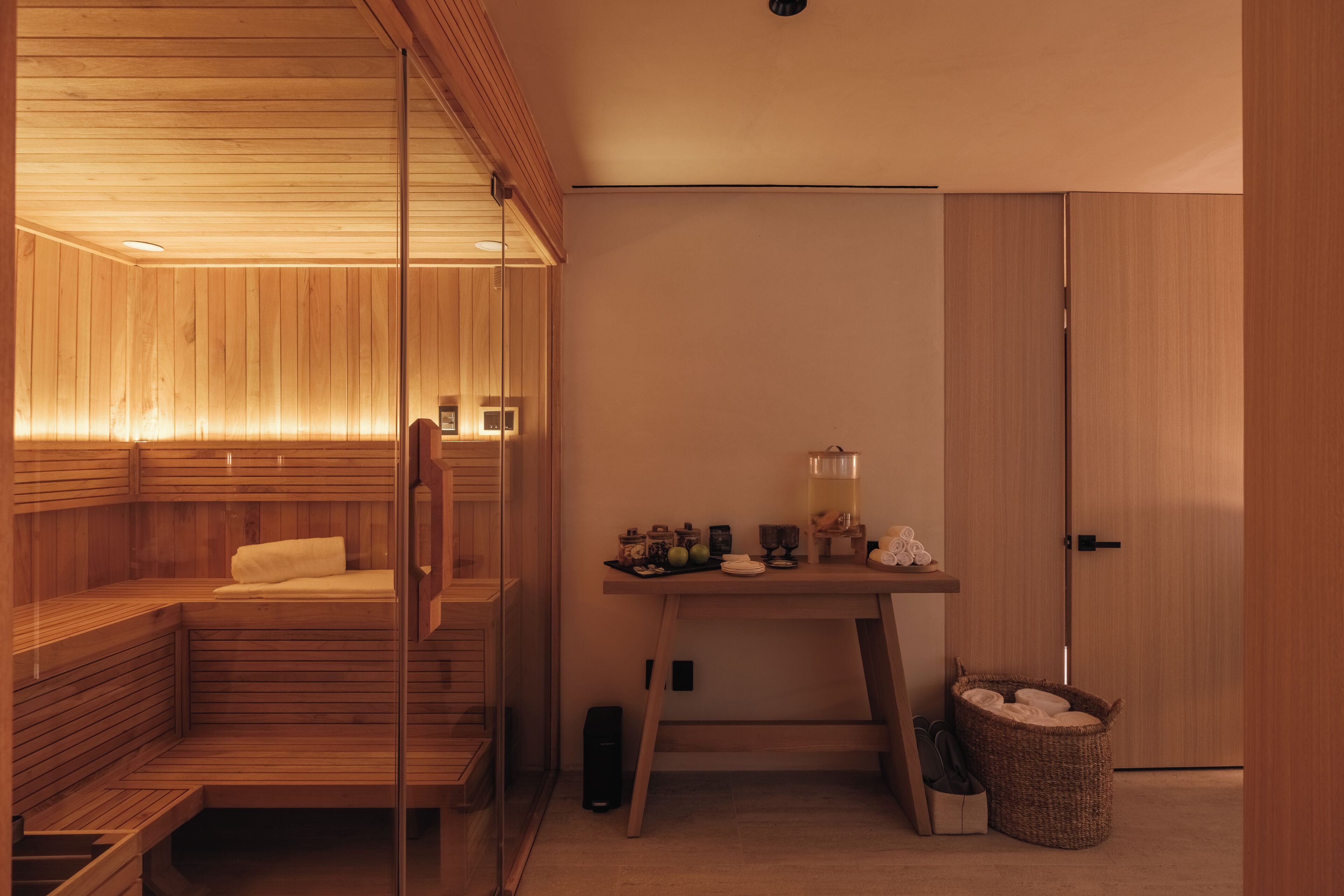 Sala de sauna con bancos de madera y una pared de vidrio, una mesita con artículos de tocador y una puerta a la derecha.