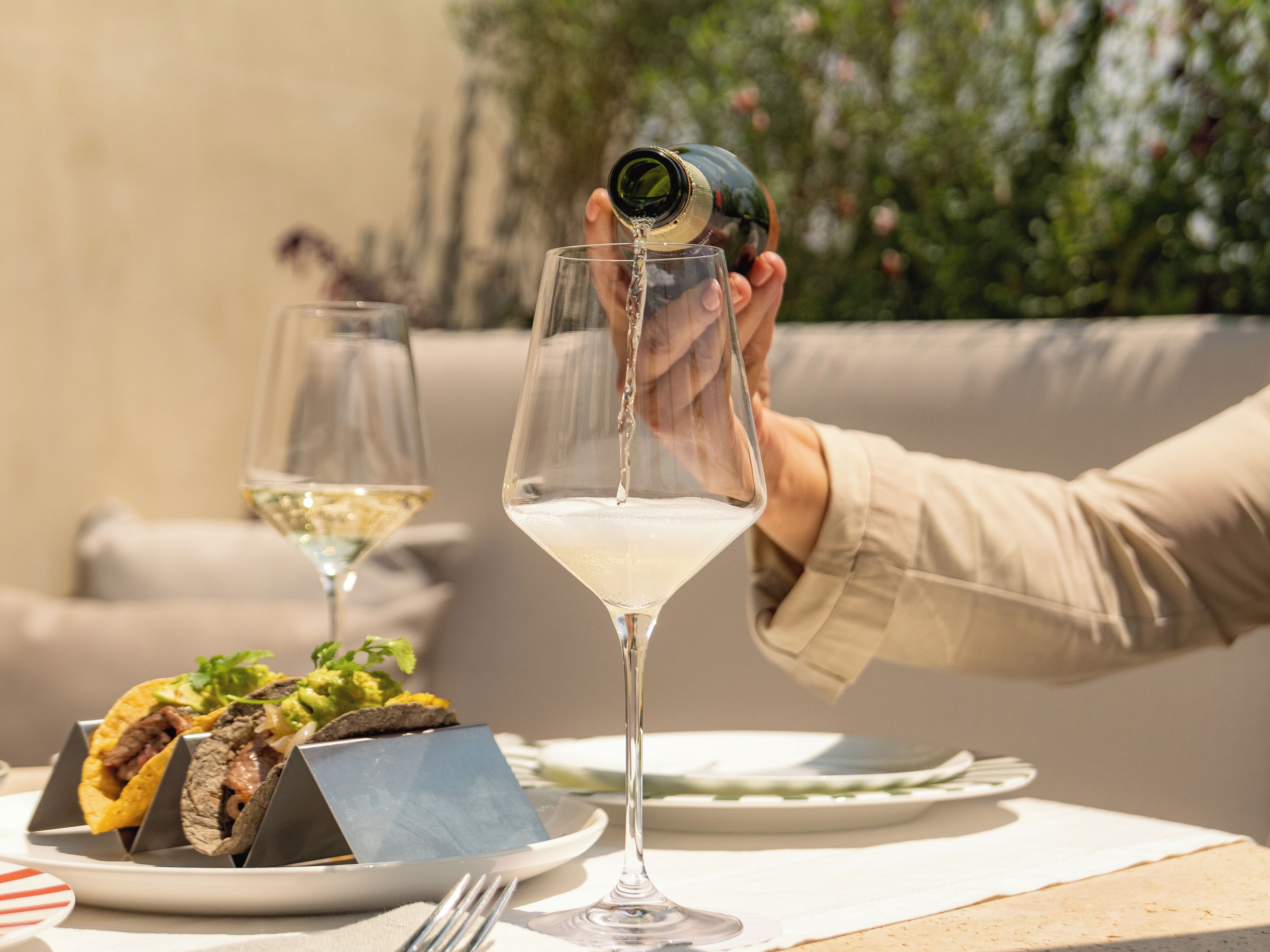 A dining table with a plate of tacos, a glass of white wine, and a bottle being poured into the glass.