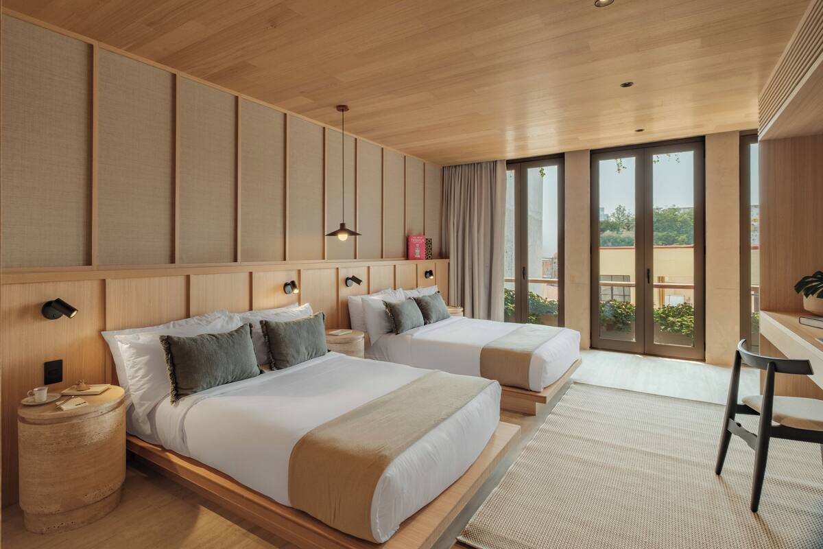 A hotel room with two beds, a desk, a chair, a waste bin, and a view of the outdoors.