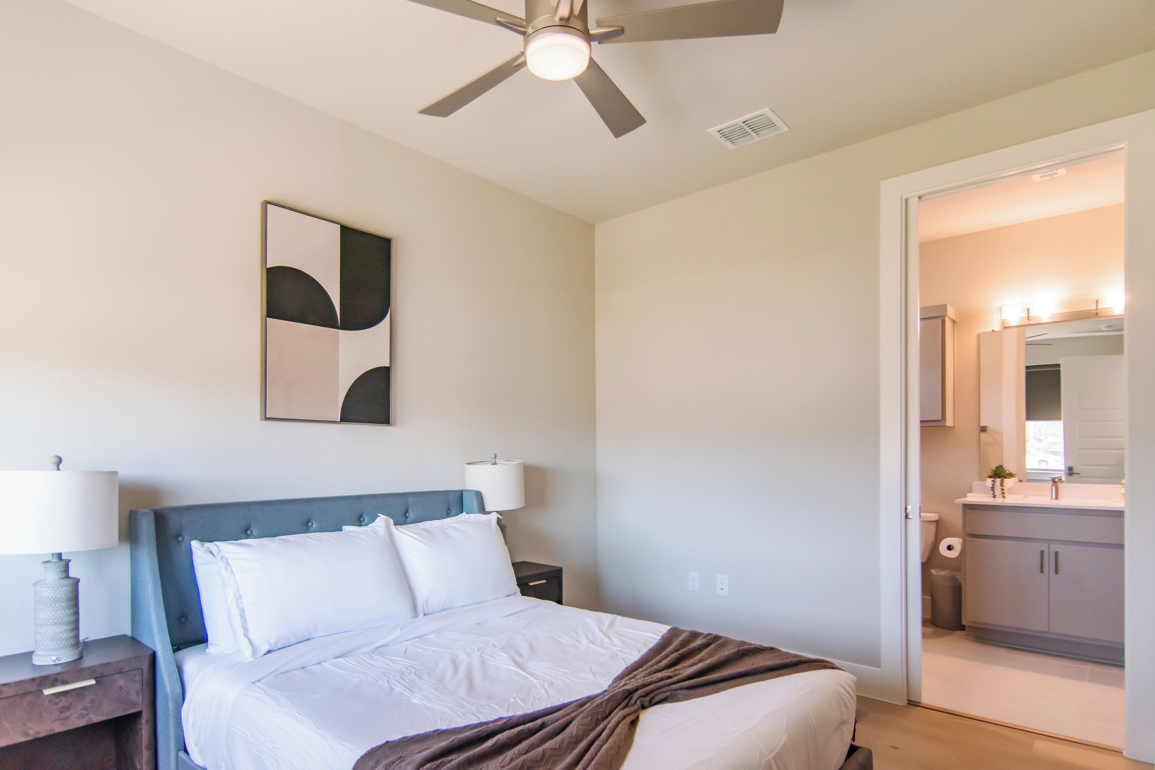 A bedroom with a bed, bedside tables, a ceiling fan, a wall-mounted abstract artwork, and a bathroom visible through an open door.