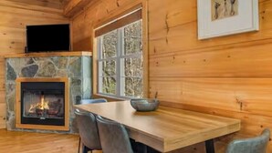 A wooden cabin interior with a stone fireplace, a dining table with chairs, and a window with a view of trees. - NEW CABIN!-Pet Friendly-Babbling Brook-Private Lake-Grill (Lake Lure)