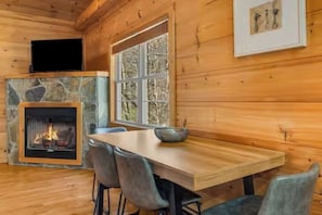 L’intérieur d’un chalet en bois, avec une cheminée en pierre, une table à manger entourée de chaises et une fenêtre donnant sur les arbres.