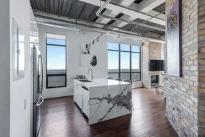 A modern kitchen with a marble island, large windows, and a brick wall.