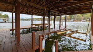A wooden dock extending over water with a covered area, wooden benches, and a view of the lake and distant buildings. - Lakefront, Dock, Kayaks, Games, Firepit, Fireplace (Littleton)