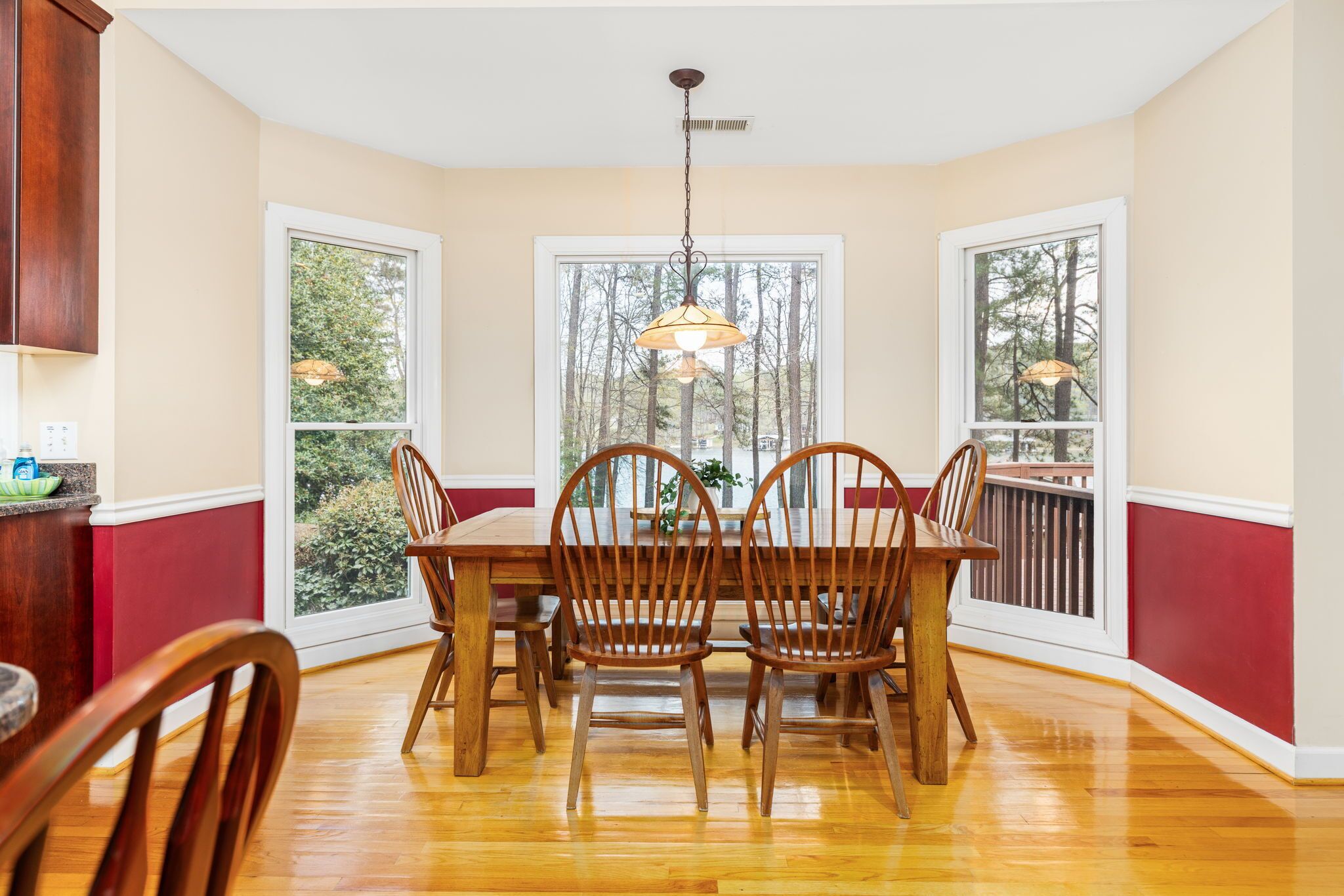 A dining area with a wooden table and chairs, large windows with a view of trees, and a hanging light fixture.