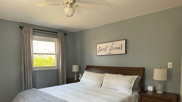 Ein Schlafzimmer mit einem Bett, zwei Nachttischen, einem Deckenventilator, einem Fenster mit Vorhängen und einem an der Wand befestigten Schild.