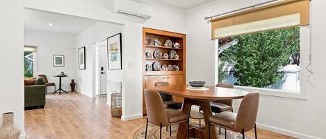 A dining area with a round table and chairs, a wooden cabinet with dishes, and a window with a view of greenery.
