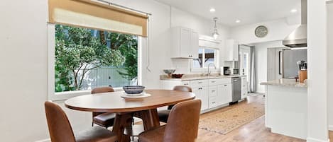 Una cocina moderna con armarios blancos, un comedor con una mesa redonda y sillas de cuero, y vista a espacios verdes.