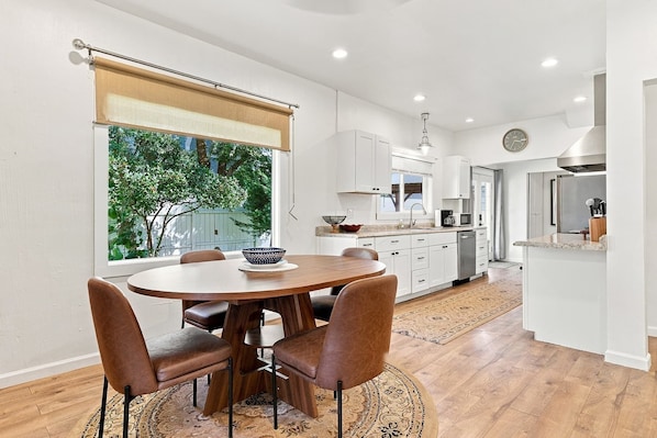 A modern kitchen with white cabinets, a dining area with a round table and leather chairs, and a view of greenery outside. - Sun Lake Retreat – Banks Lake Getaway (Coulee City)