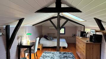 A loft bedroom with a sloped ceiling, a bed, a desk with a lamp, a dresser, and a window.