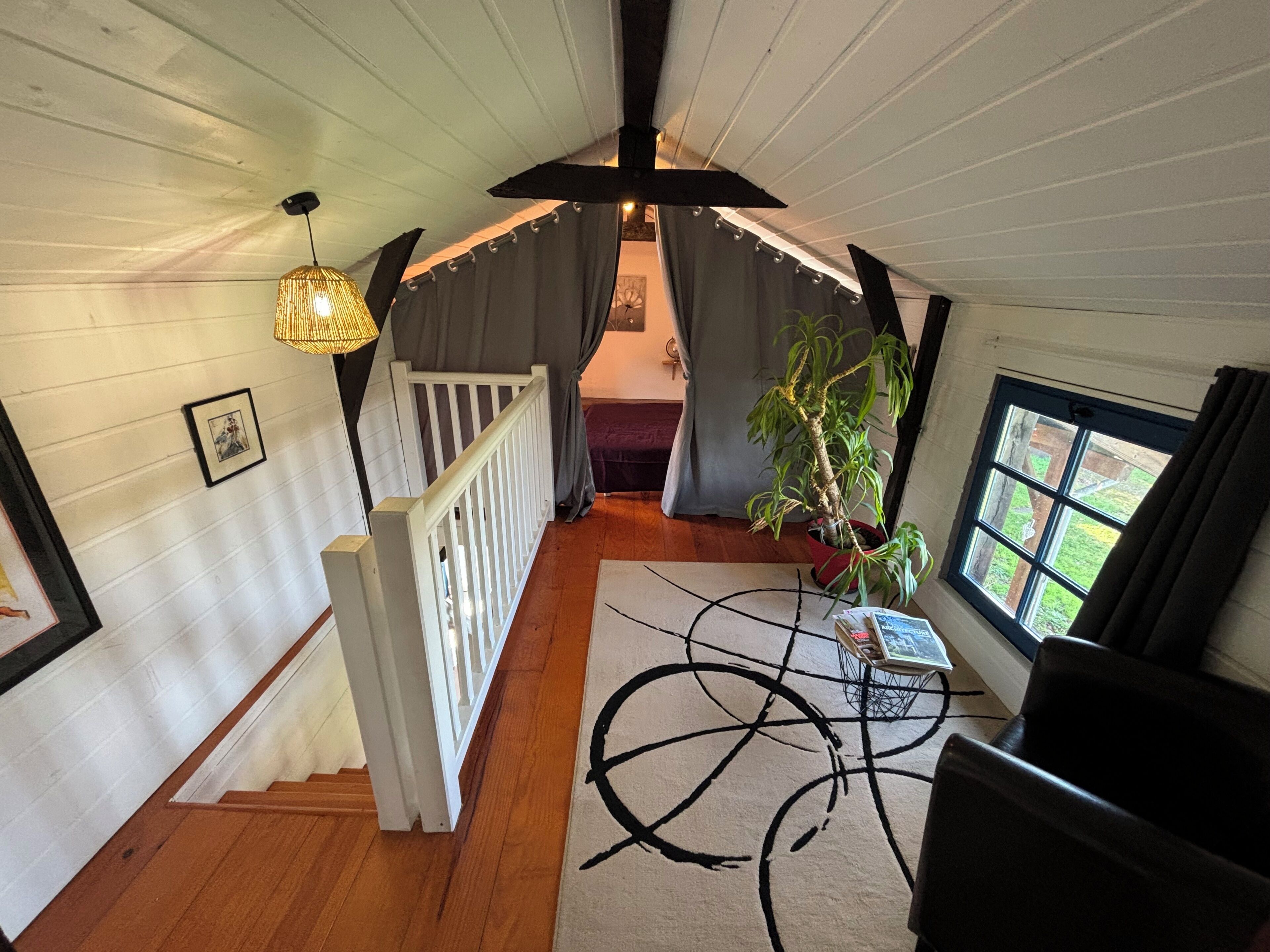 A room with a wooden floor, a white railing, a staircase, a potted plant, a table with books, and a window with curtains.
