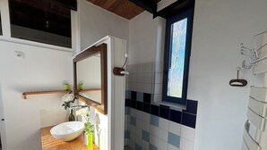 Ein Badezimmer mit einem Holzkosmetikschrank, einem weißen Waschbecken, einem Spiegel, einem Fenster und einem gefliesten Duschbereich.
