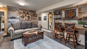 A cozy living room with a sofa, coffee table, and two bar stools. A dartboard is mounted on the wall.