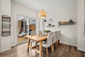 Ein Essbereich mit einem Holztisch, grau gepolsterten Stühlen, einer Hängeleuchte und Blick in eine Küche.