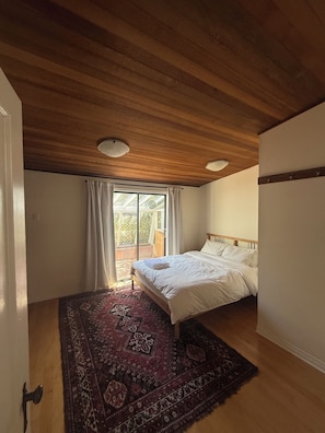A bedroom with a wooden ceiling, a bed with white linens, a patterned rug, and a large window with curtains.