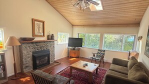 A living room with a stone fireplace, a ceiling fan, a television, a sofa, a coffee table, and a patterned rug. - 2 Bedroom Semi-Waterfront Cottage in Vesuvius, Salt Spring Island (Salt Spring Island)