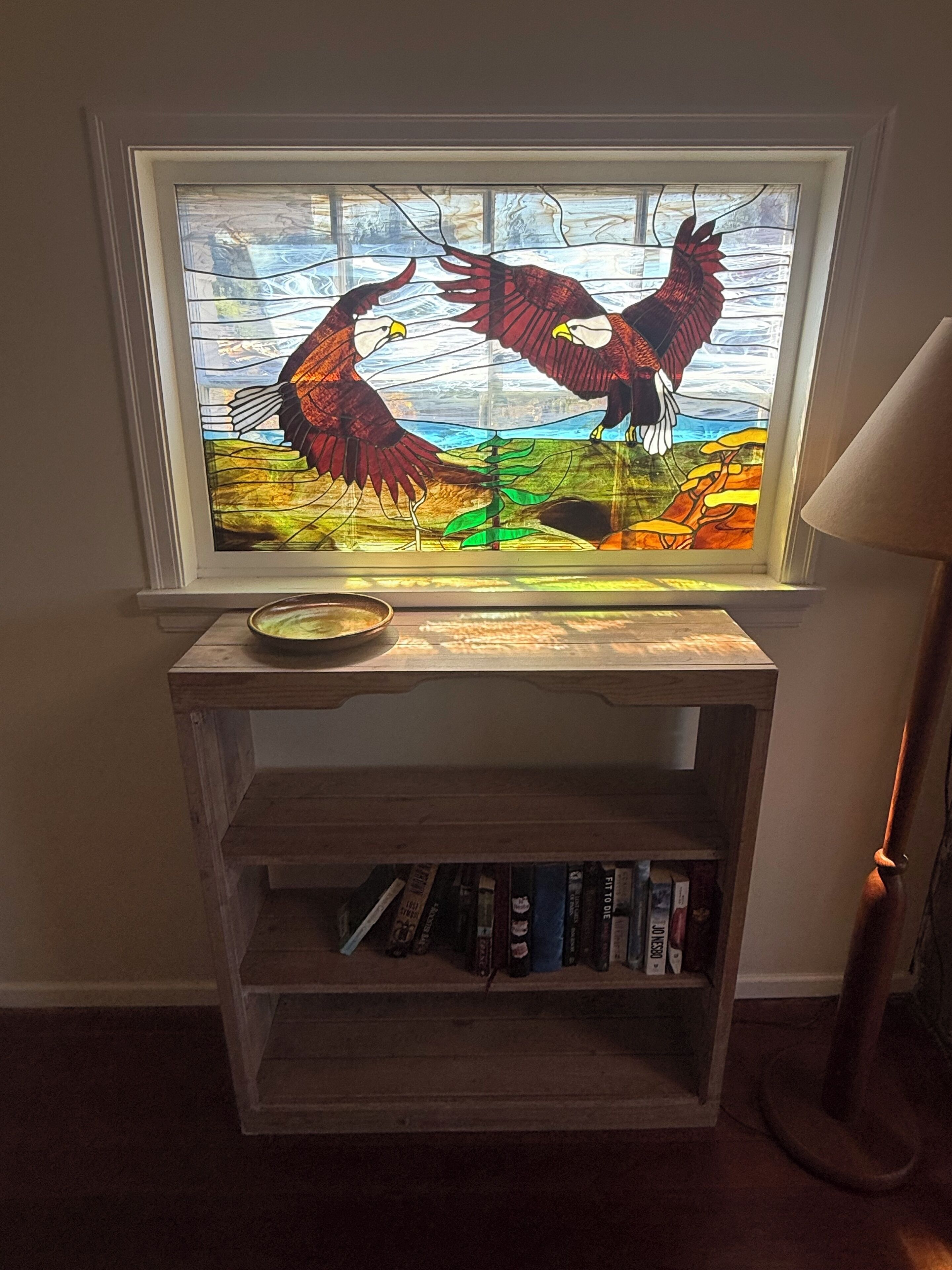 Una ventana de vitral con dos águilas, una estantería de madera con libros y una lámpara.
