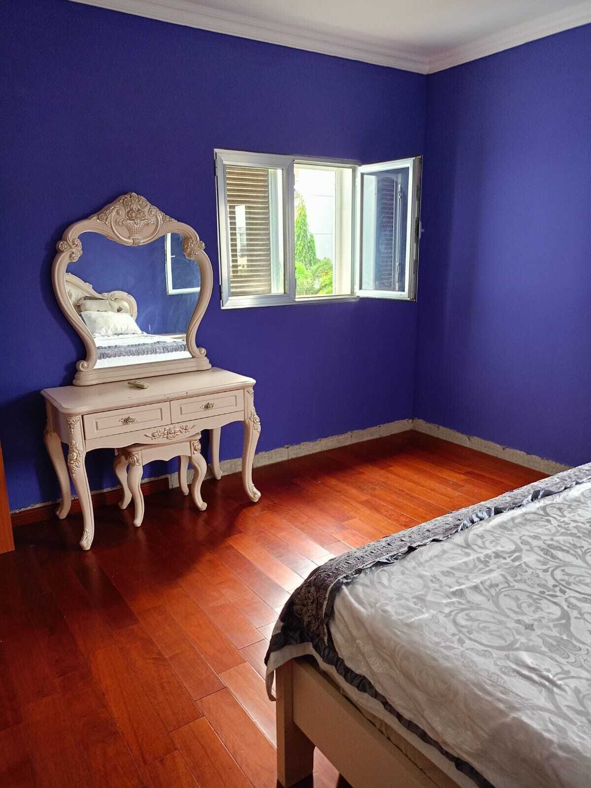 A bedroom with a large bed, a dresser with drawers, a mirror, and a window with shutters.