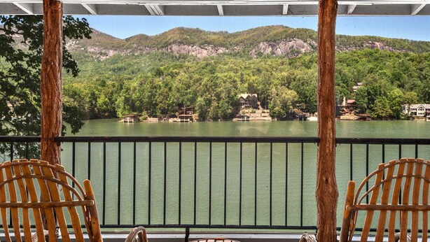 A covered outdoor area with wooden chairs and a table, overlooking a lake with houses and mountains in the background.