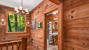 A wooden cabin interior with a dining area, a bathroom with a sink and mirror, and a window with a view of trees.