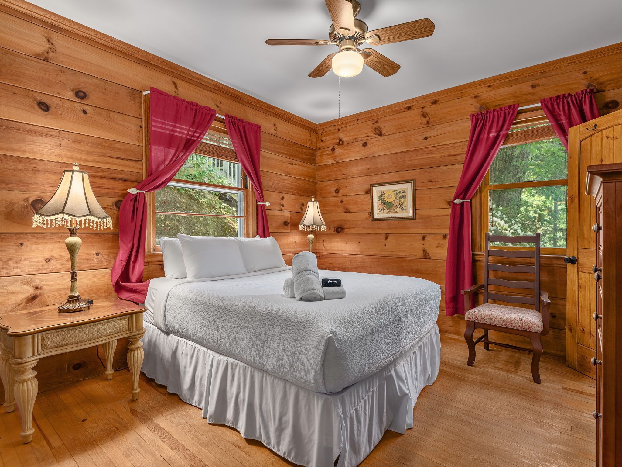 Un dormitorio de cabaña de madera con cama, mesita de noche, lámpara, silla y ventilador de techo.