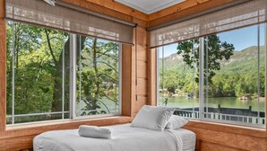 A wooden room with a bed, large windows, and a view of a lake and forest.