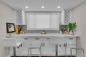 Une cuisine moderne avec des meubles blancs, une grande fenêtre équipée de stores et trois tabourets de bar.