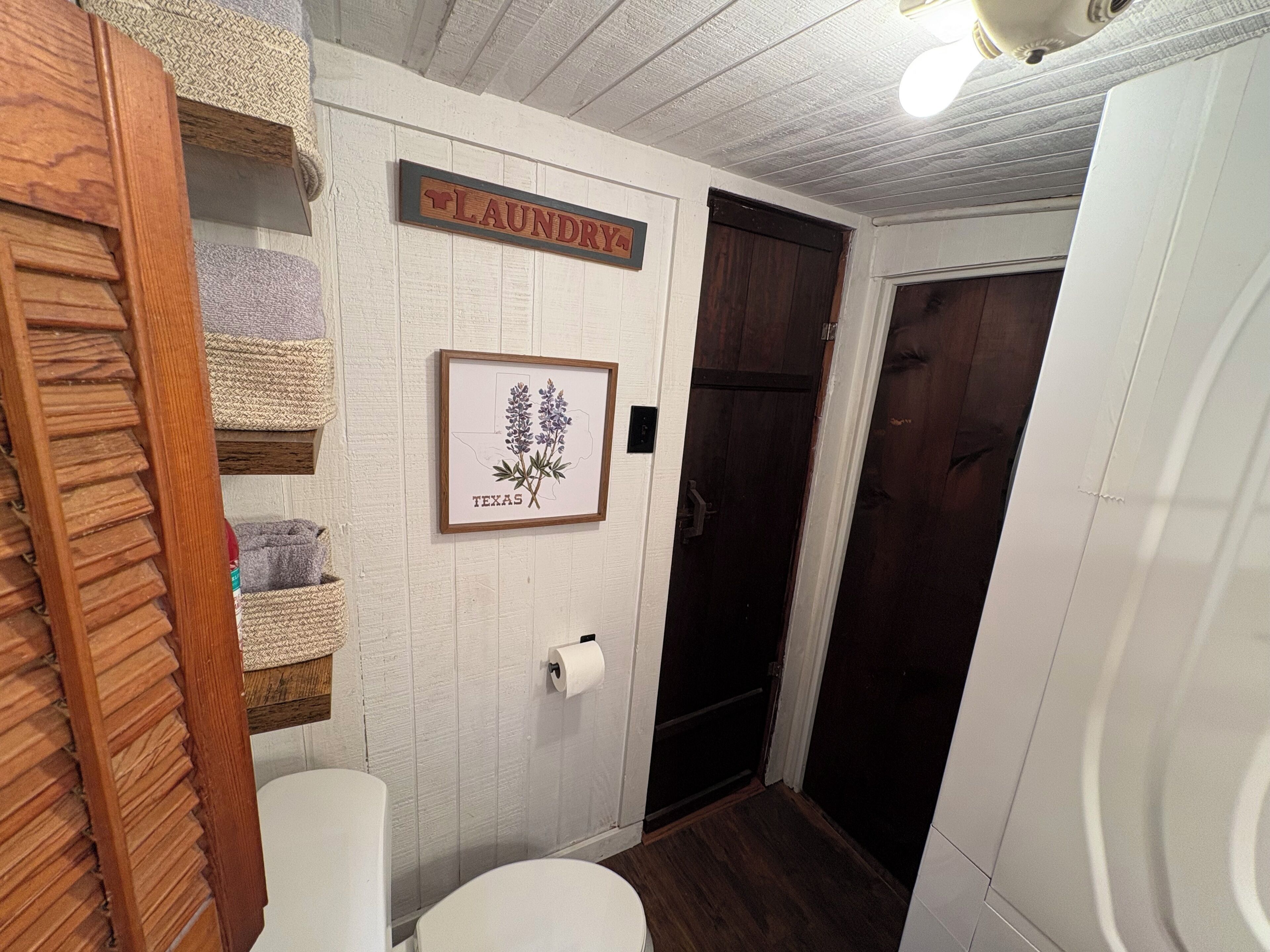 A bathroom with a wooden shelf, a framed picture of a plant, a toilet, and a door.