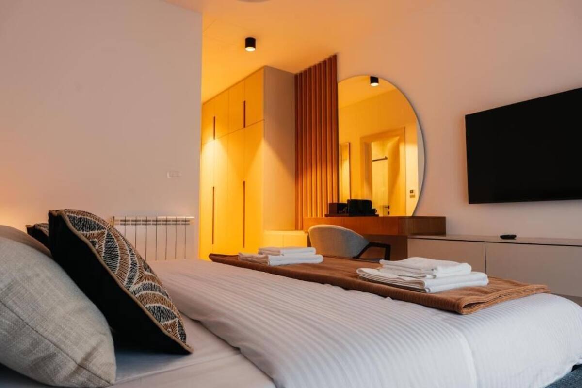Habitación de hotel con cama, mesita de madera con toallas, una silla, escritorio, espejo y televisor.
