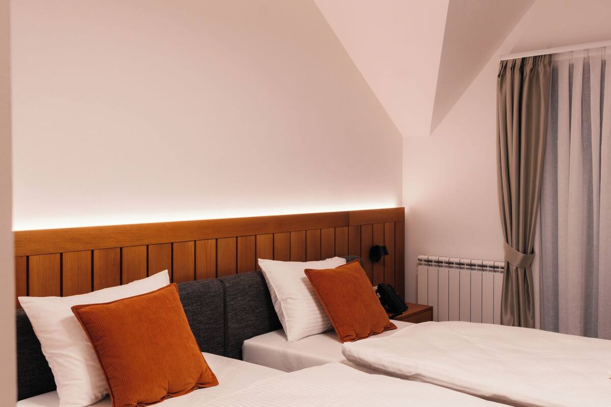 Habitación de hotel con dos camas, cabecero de madera, una lámpara en el techo, un radiador y una ventana con cortinas.