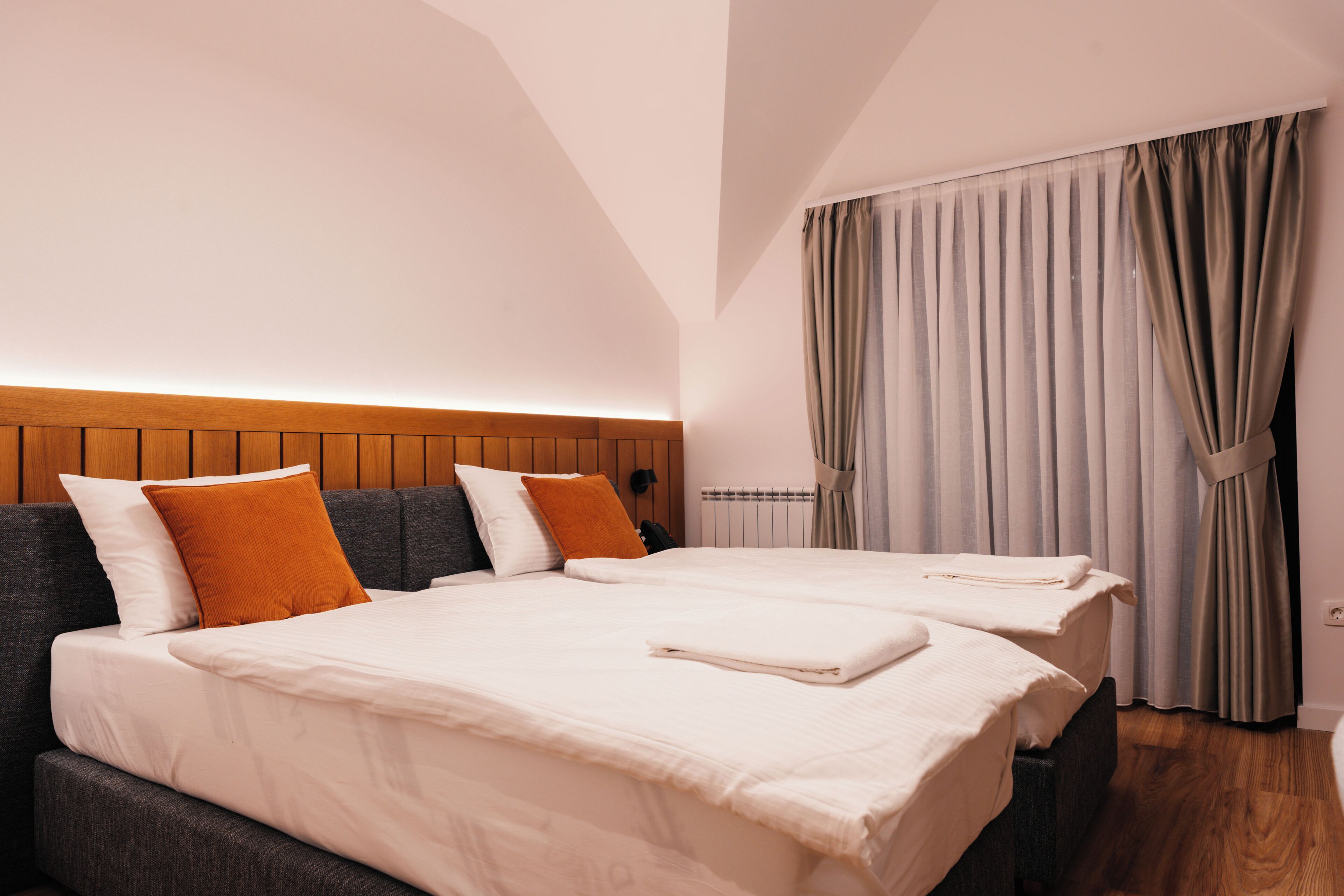 A hotel room with two beds, a wooden headboard, a window with curtains, and a radiator.