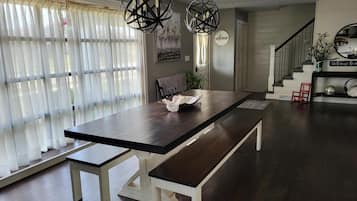 A spacious dining area with a long table, two hanging light fixtures, and a staircase leading to an upper level.