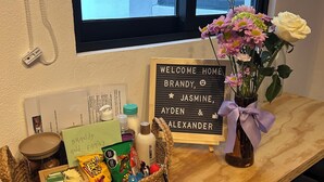 Una habitación con un escritorio de madera, un cartel de bienvenida, un jarrón con flores y una cesta de aperitivos.