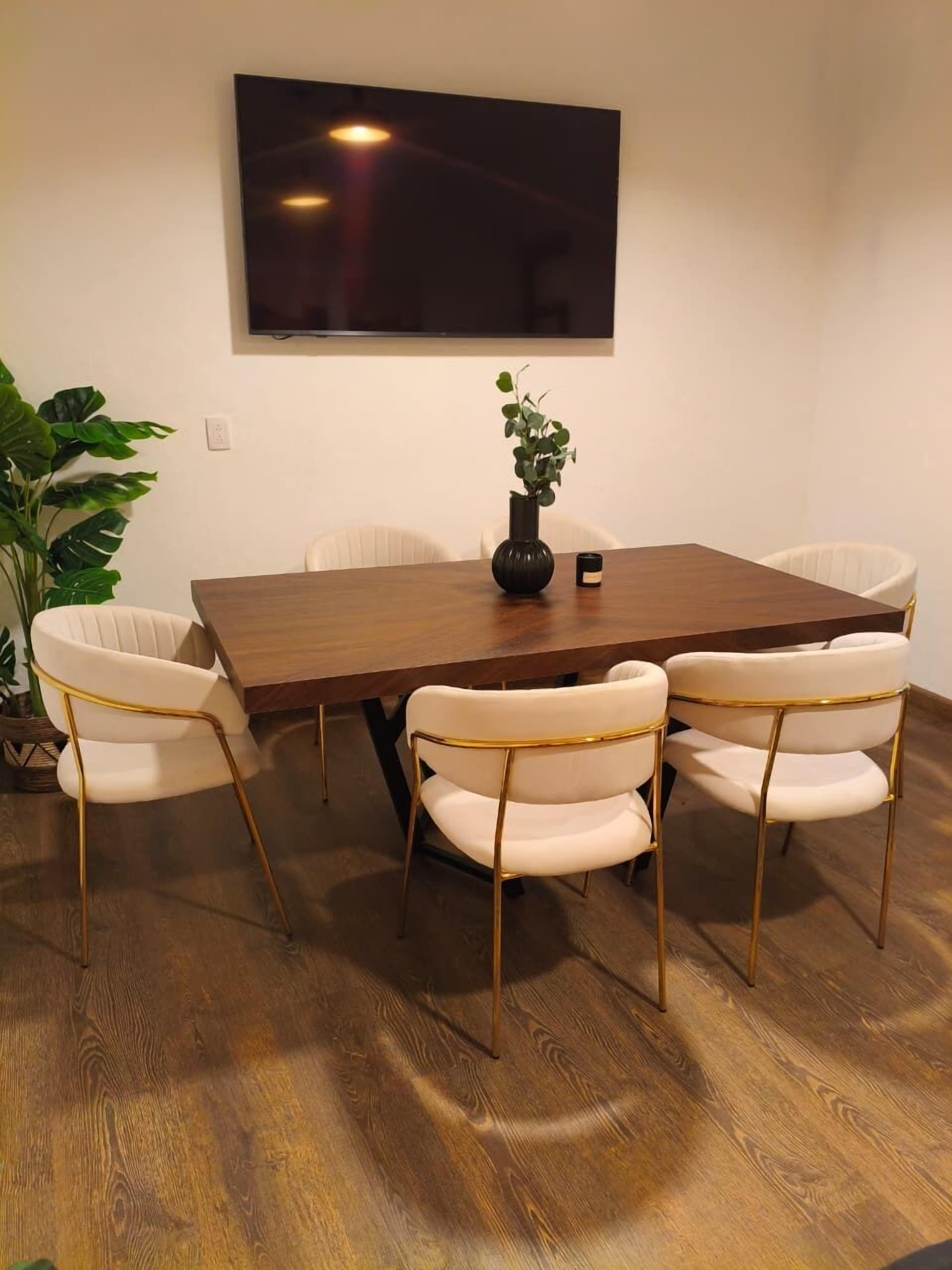 A dining area with a wooden table, four white chairs with gold legs, a potted plant, and a flat-screen TV mounted on the wall.