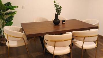A dining area with a wooden table, four white chairs with gold legs, a potted plant, and a flat-screen TV mounted on the wall.
