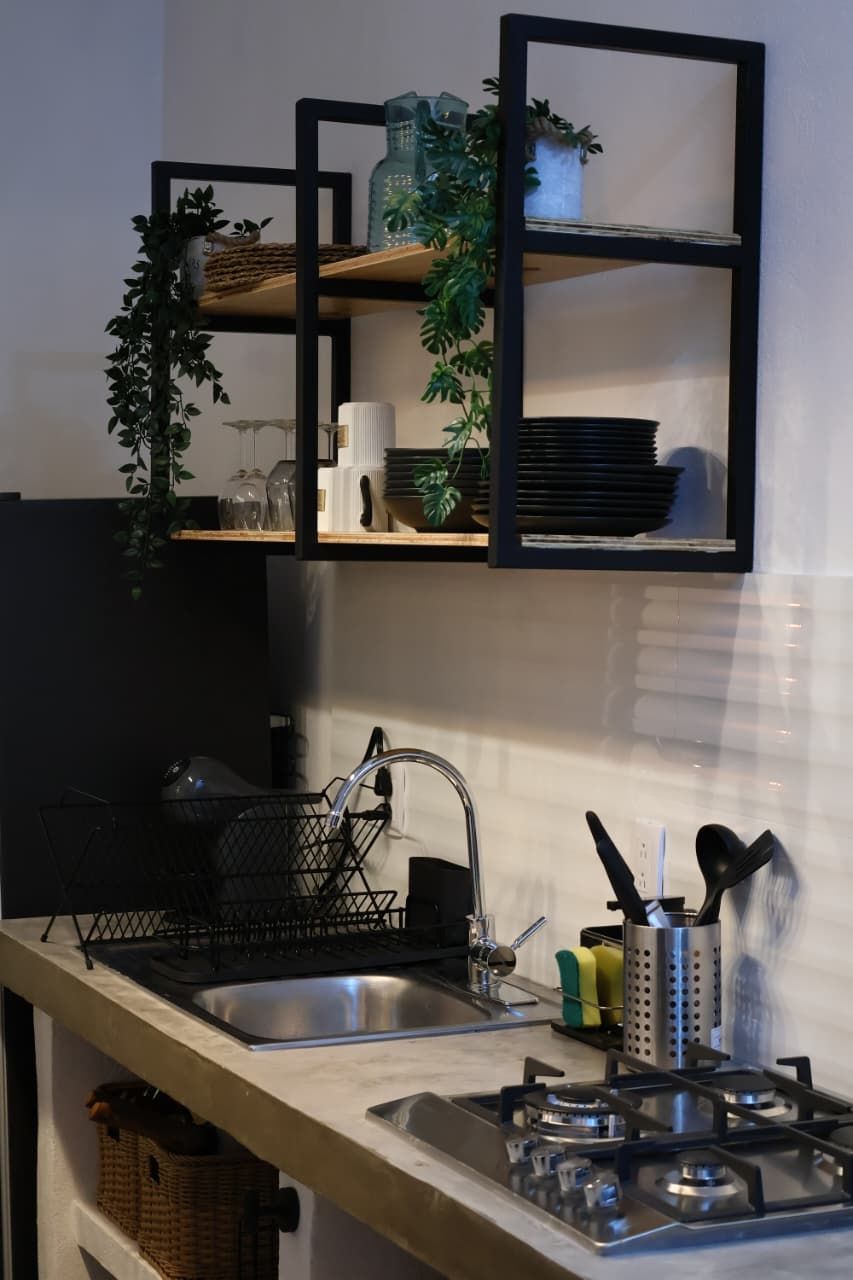 Une cuisine équipée d’une cuisinière à gaz intégrée, d’un évier et d’une étagère en métal noir au-dessus du plan de travail, où sont rangés les ustensiles de cuisine et des plantes.