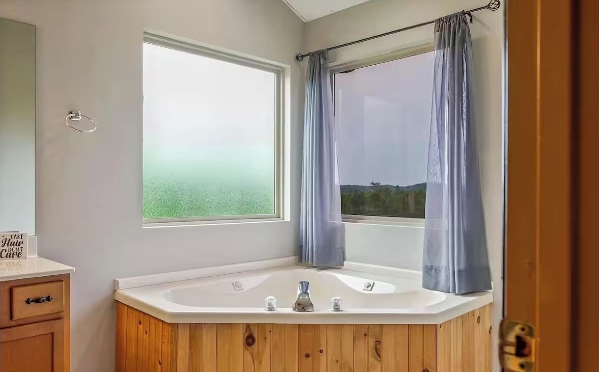 Une salle de bain avec un meuble-lavabo en bois, une grande fenêtre avec des rideaux et une vue sur la verdure à l’extérieur.