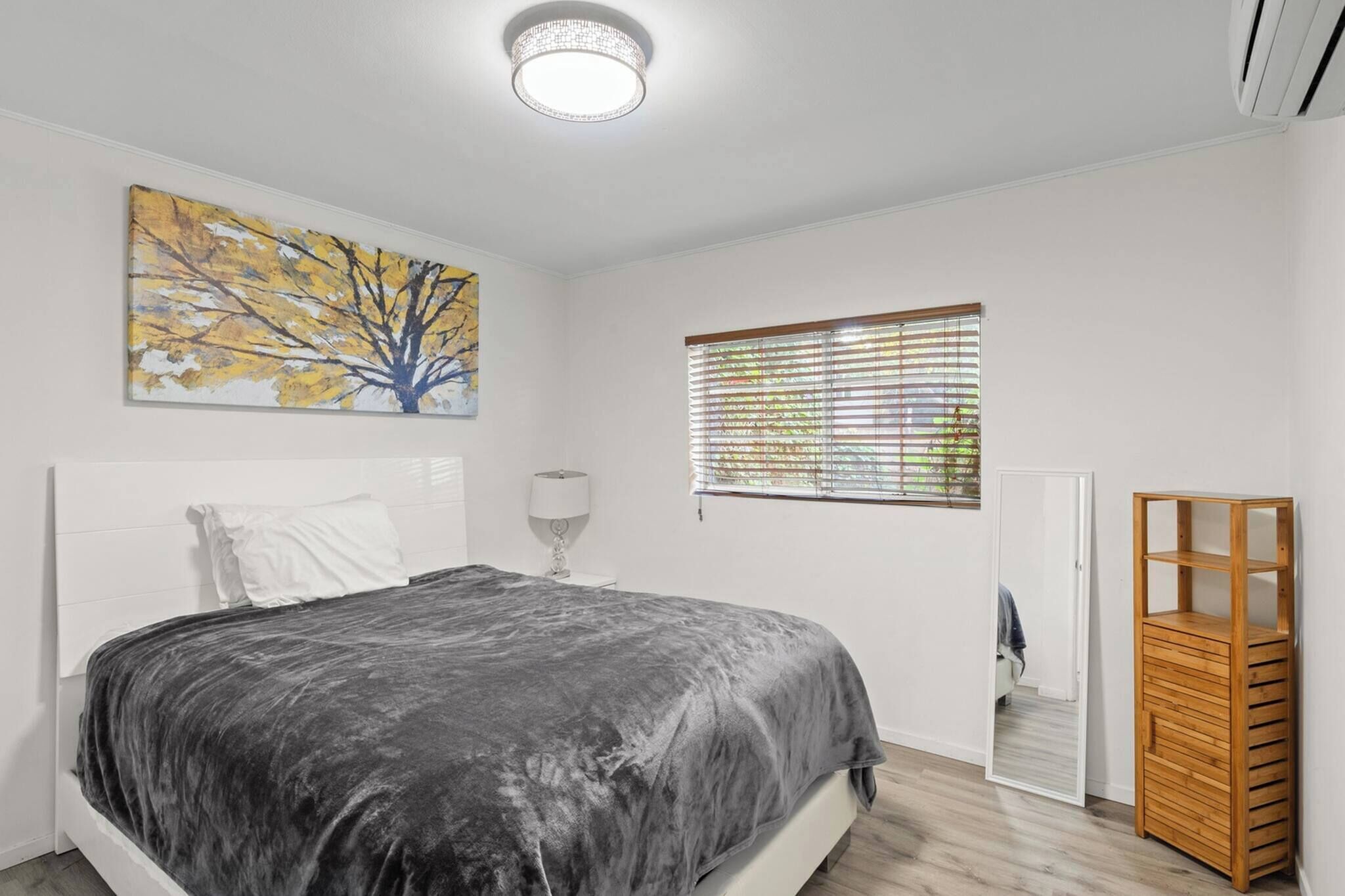 A bedroom with a bed, a bedside table, a lamp, a window with blinds, a door, and a wooden storage unit.