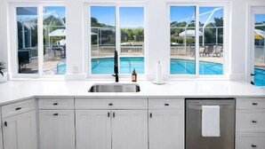 Una cocina con armarios blancos, un lavamanos de acero inoxidable y vistas a una piscina a través de amplios ventanales.
